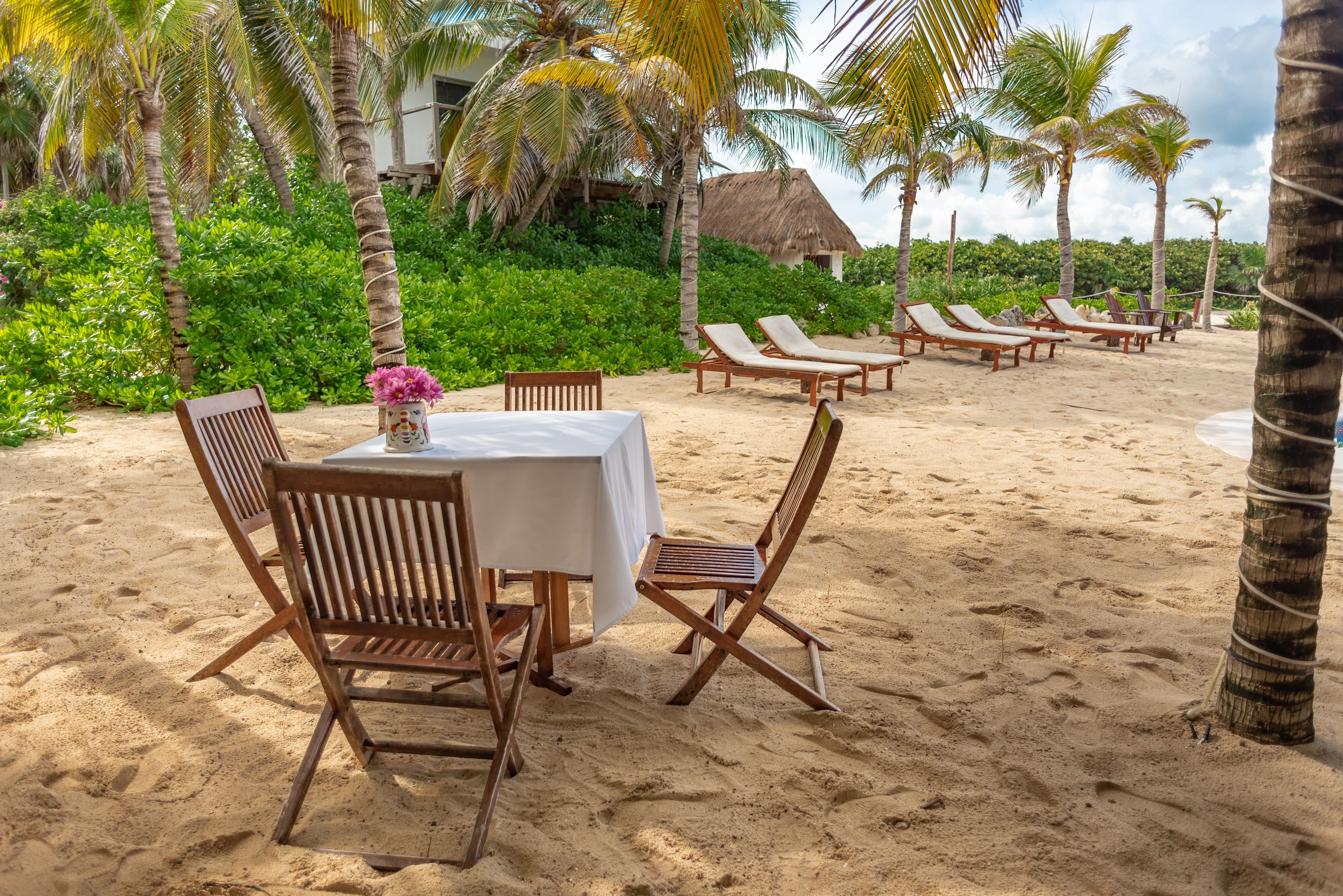 A beachside dining area with wooden chairs and a table set for two, surrounded by palm trees and a thatched-roof building in the background.