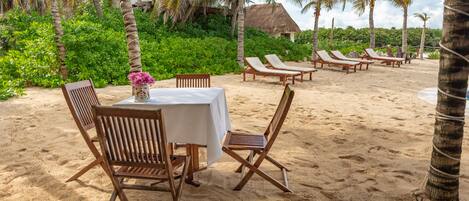A beachside dining area with wooden chairs and a table set for two, surrounded by palm trees and a thatched-roof building in the background.