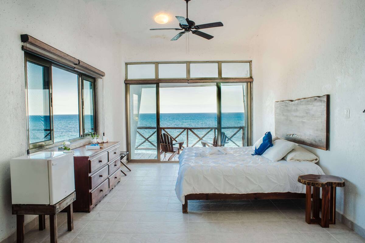 A bedroom with a large bed, a dresser, a ceiling fan, and a view of the ocean through a glass door.