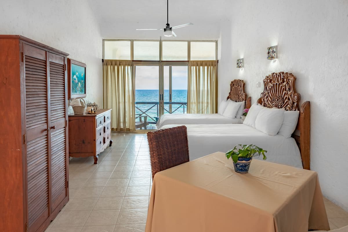 A hotel room with a bed, a table with a plant, a wardrobe, a dresser, and a view of the sea through large windows.