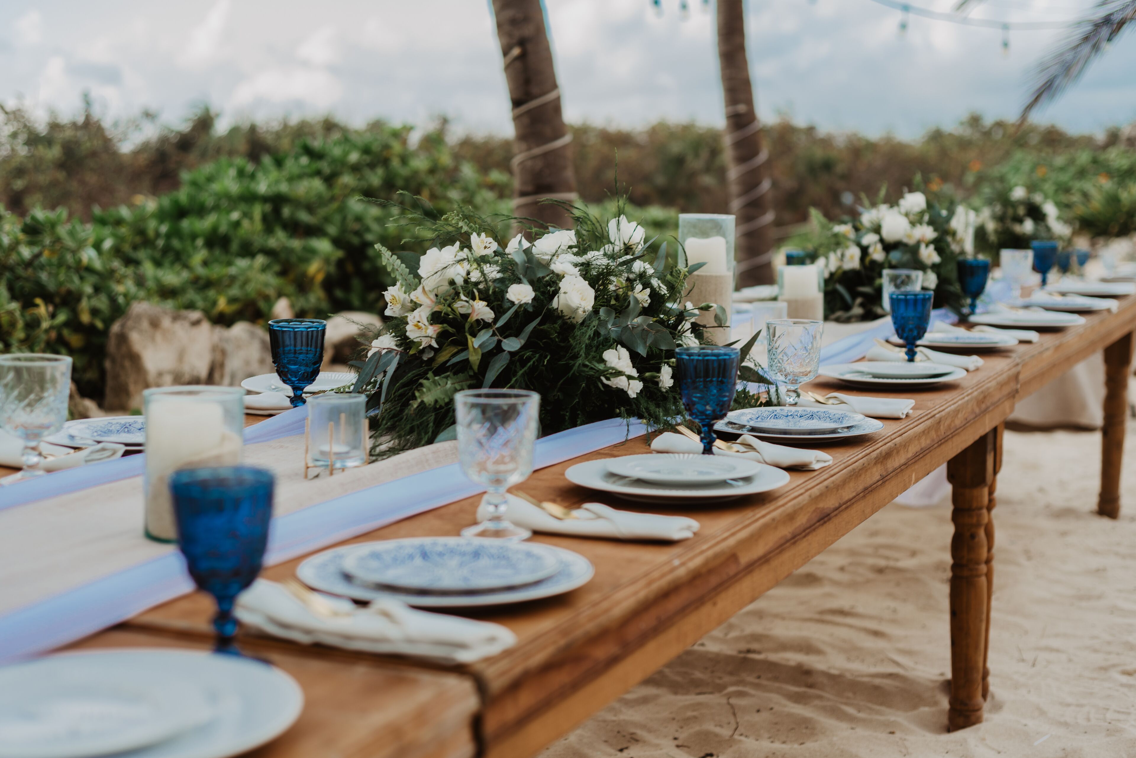 Une longue table en bois, dressée sur une plage de sable, est ornée d’assiettes, de verres et de centres de table floraux.