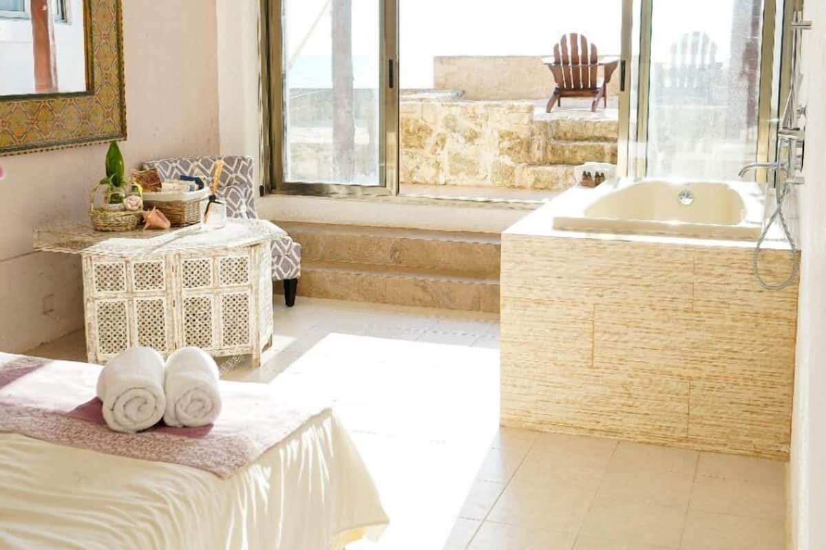 A spacious bedroom with a large bed, a bedside table, a small table with decorative items, and a view of a balcony with a chair.