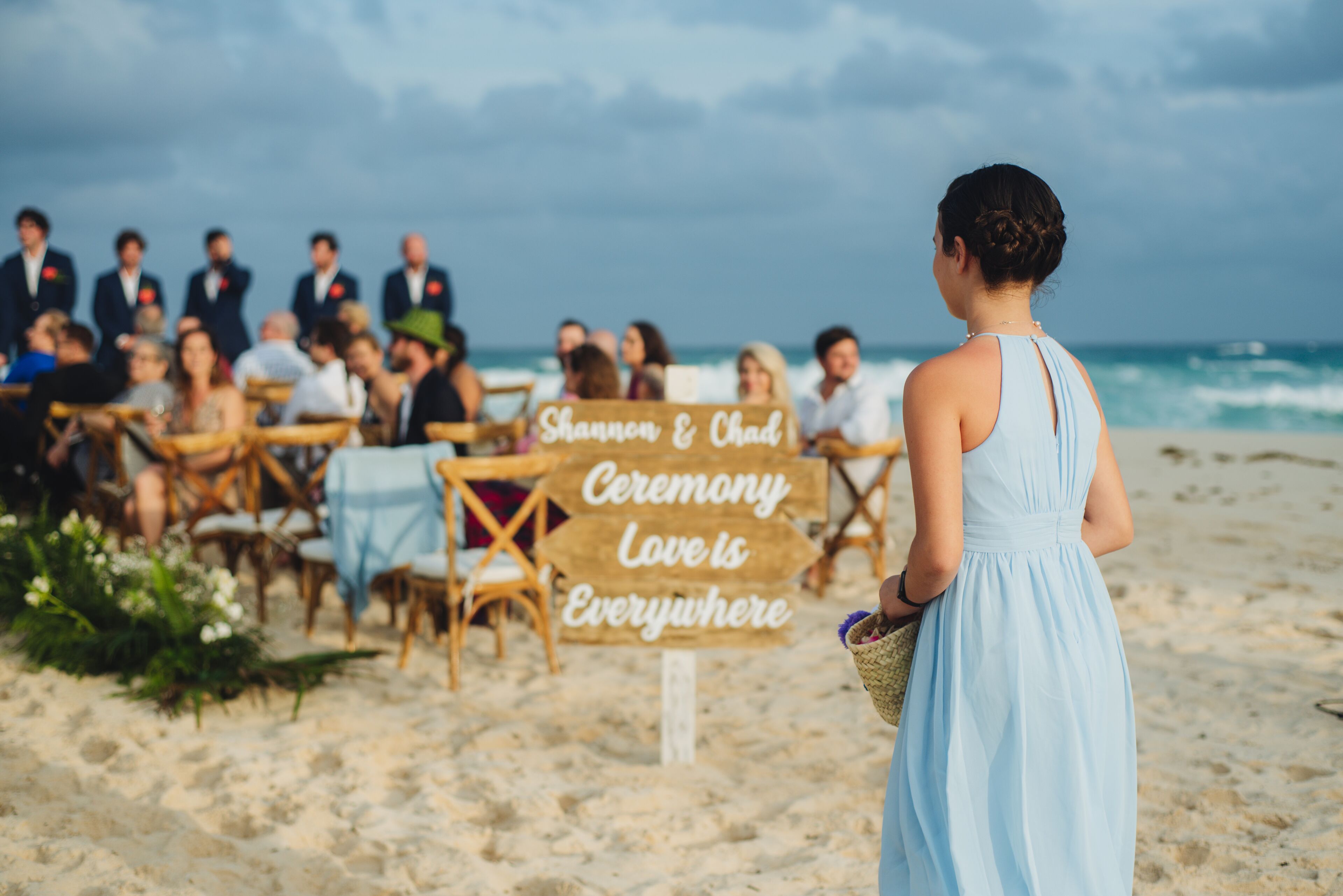 Una ceremonia de boda en la playa, con invitados sentados en mesas y un cartel que dice 'Shannon & Chad Ceremony Love is Everywhere'.