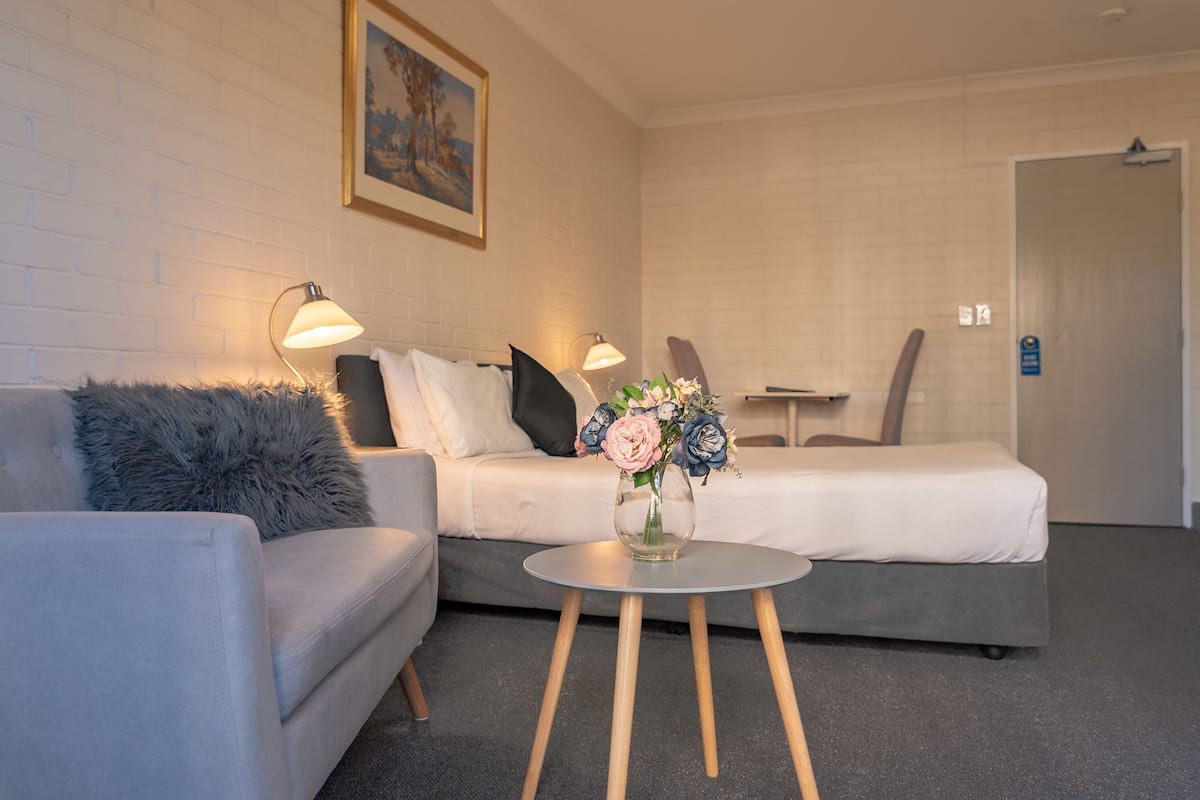 A modern hotel room with a grey sofa, a small round table with a vase of flowers, and a bed with a white headboard.