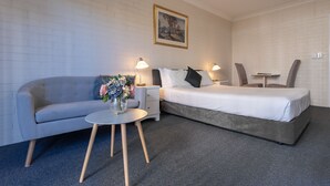 A modern hotel room with a grey sofa, a small round table with a vase of flowers, a bed with white bedding, and two bedside lamps. - Benson Court Motel (Toowong)