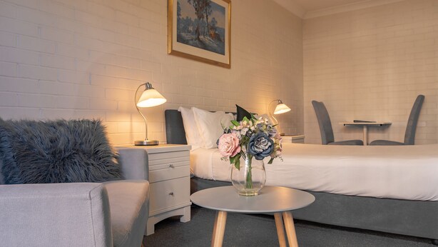 A hotel room with a bed, a chair, a small table with a vase of flowers, and two bedside lamps. - Benson Court Motel (Toowong)