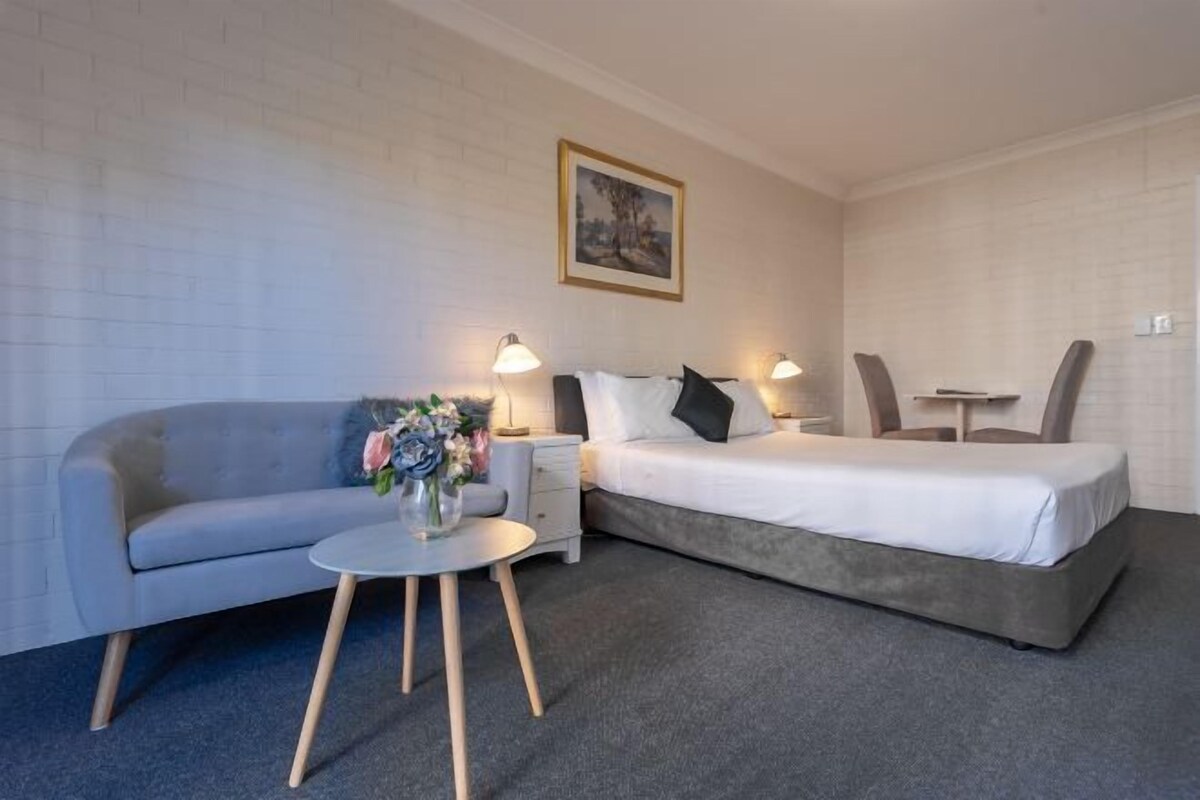A hotel room with a grey sofa, a single bed, a small round table, and two bedside lamps.