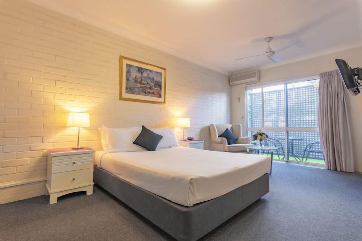 A hotel room with a large bed, two bedside tables with lamps, a chair, a balcony with a table and chairs, and a ceiling fan.