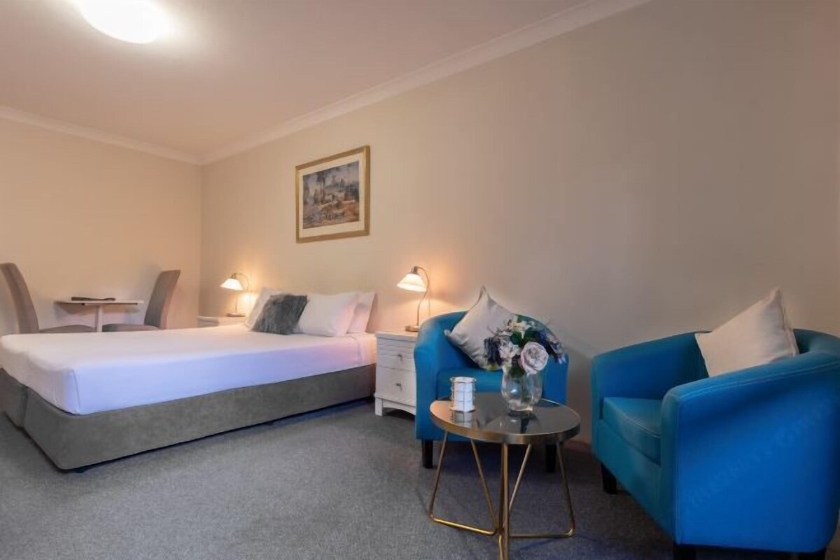 A hotel room with a bed, a blue armchair, a small table with a vase, and a nightstand with a lamp.