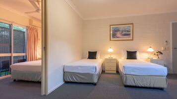 A hotel room with two beds, a ceiling fan, a window with curtains, a bedside table with a lamp, and a framed picture on the wall.