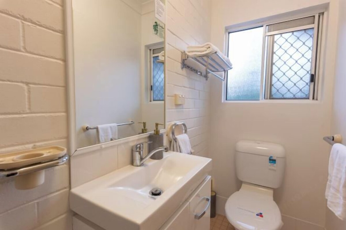 A bathroom with a white sink, a toilet, a mirror, and a window with a metal screen.
