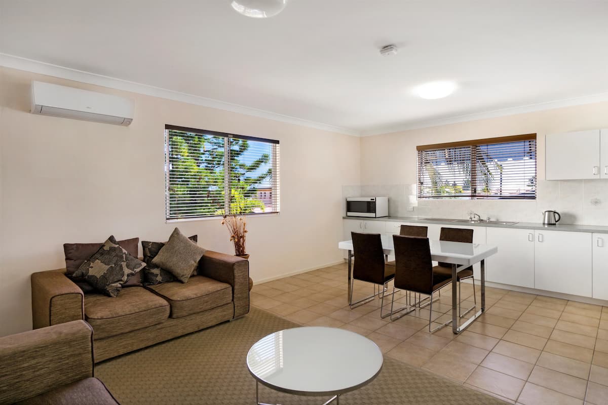 A compact living space with a sofa, dining table, chairs, a kitchen area with a microwave, and a window with blinds.