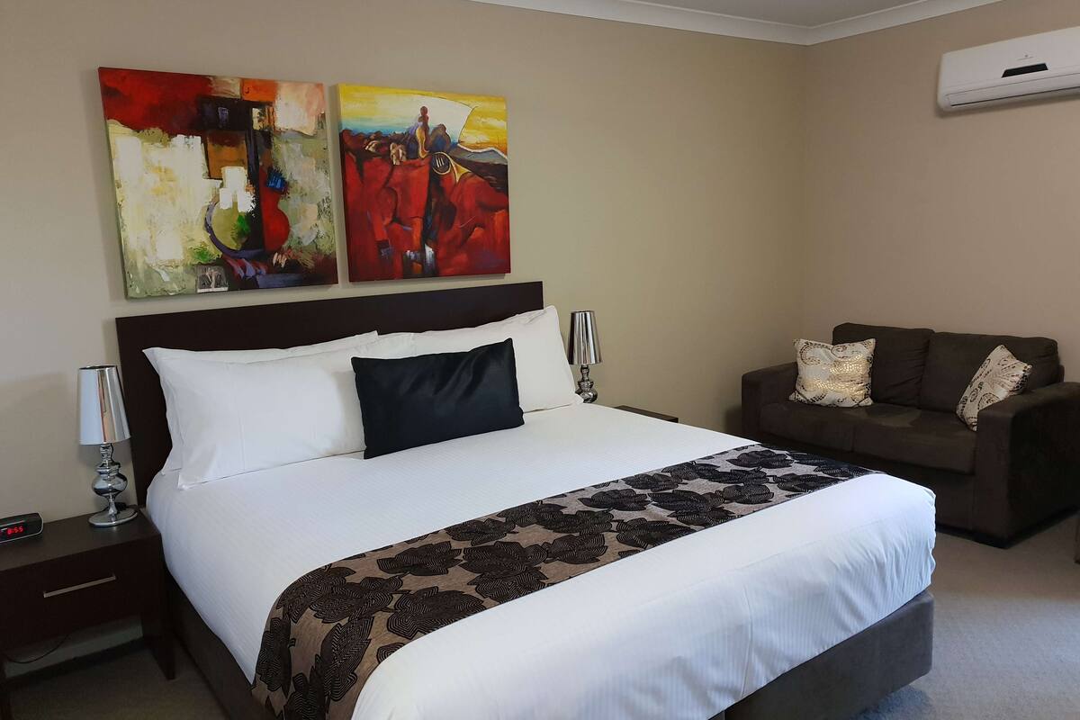 A hotel room with a large bed, a bedside table with a lamp, a brown sofa, and two abstract paintings on the wall.