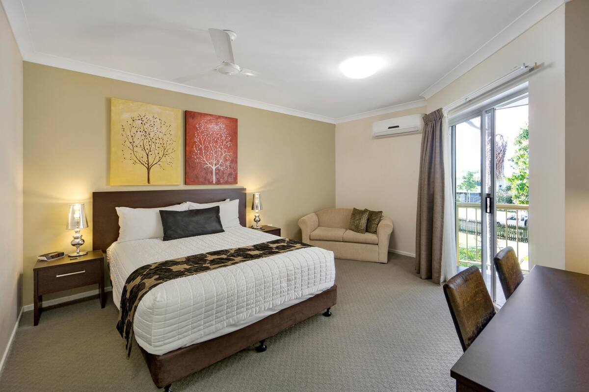 A hotel room with a large bed, a bedside table with a lamp, a sofa, and a dining table.