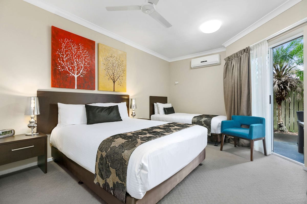 A hotel room with two beds, a ceiling fan, artwork on the wall, and a view of the outdoors.