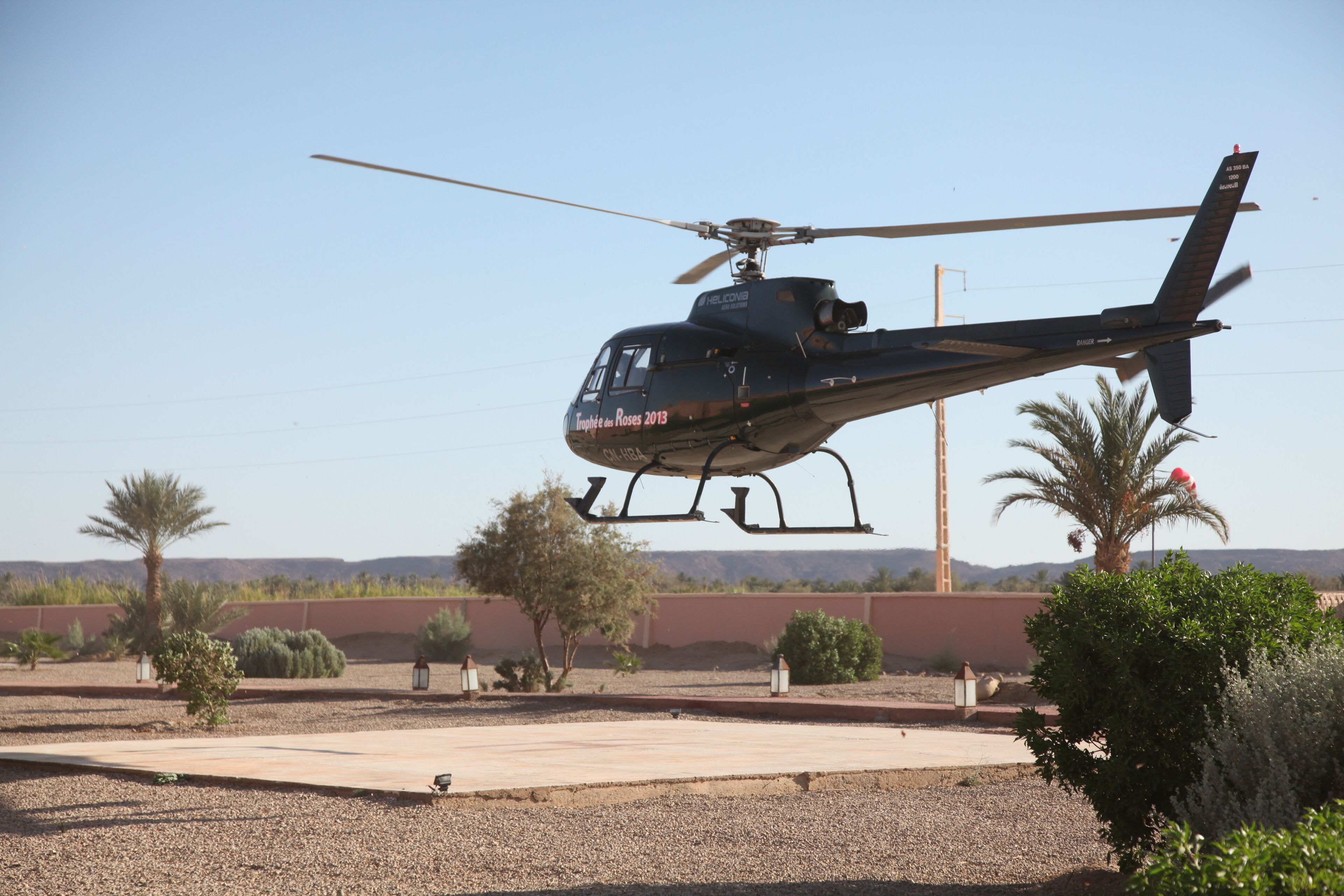Un helicóptero con la leyenda “Trophee de Roses 2013” en su lateral, suspendido sobre una zona pavimentada con palmeras y arbustos.