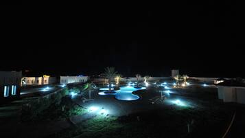 Una vista nocturna de un resort con piscinas iluminadas, palmeras y edificios modernos.