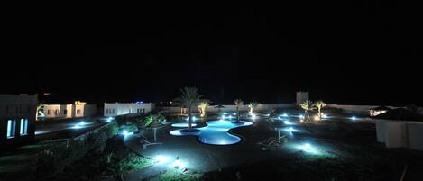 Una vista nocturna de un resort con piscinas iluminadas, palmeras y edificios modernos.