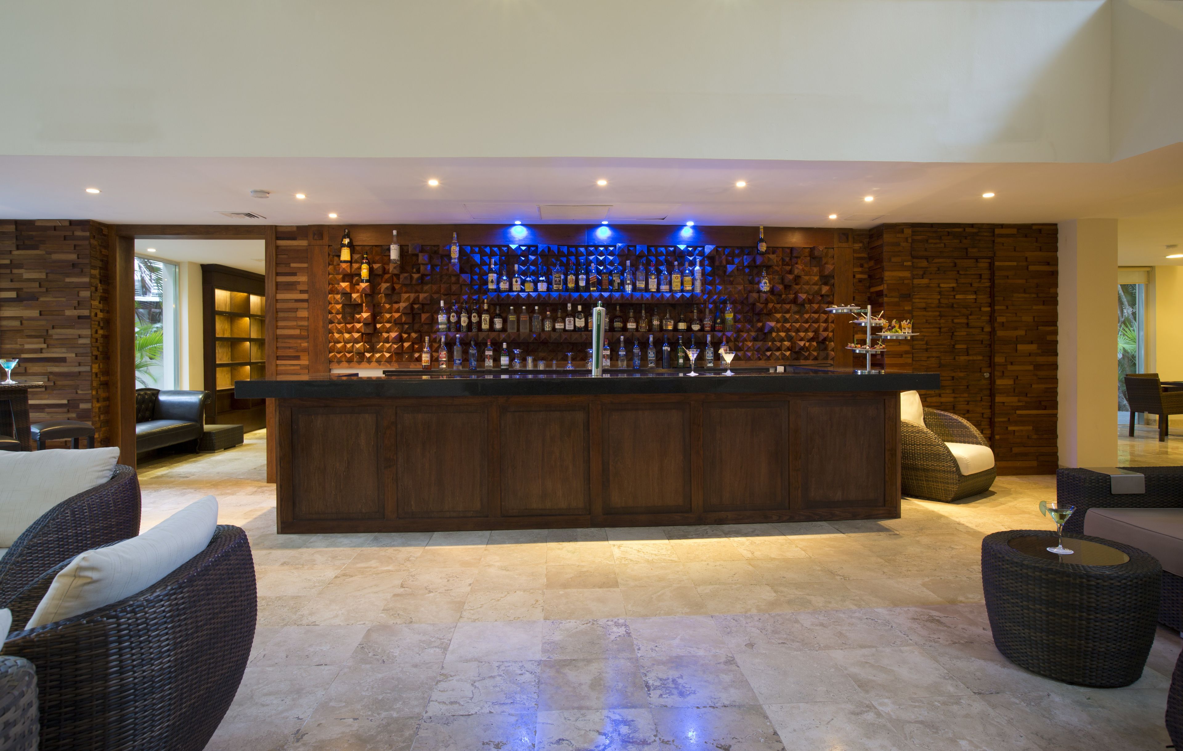 A modern bar area with a wooden bar counter, a variety of bottles, and a seating area with wicker furniture.