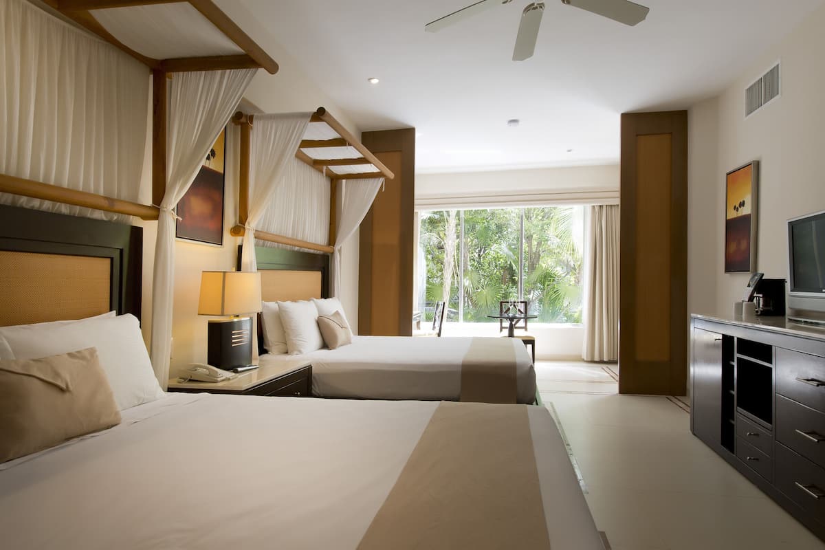 A hotel room with two beds, a desk with a lamp, a TV, and a large window with a view of greenery.