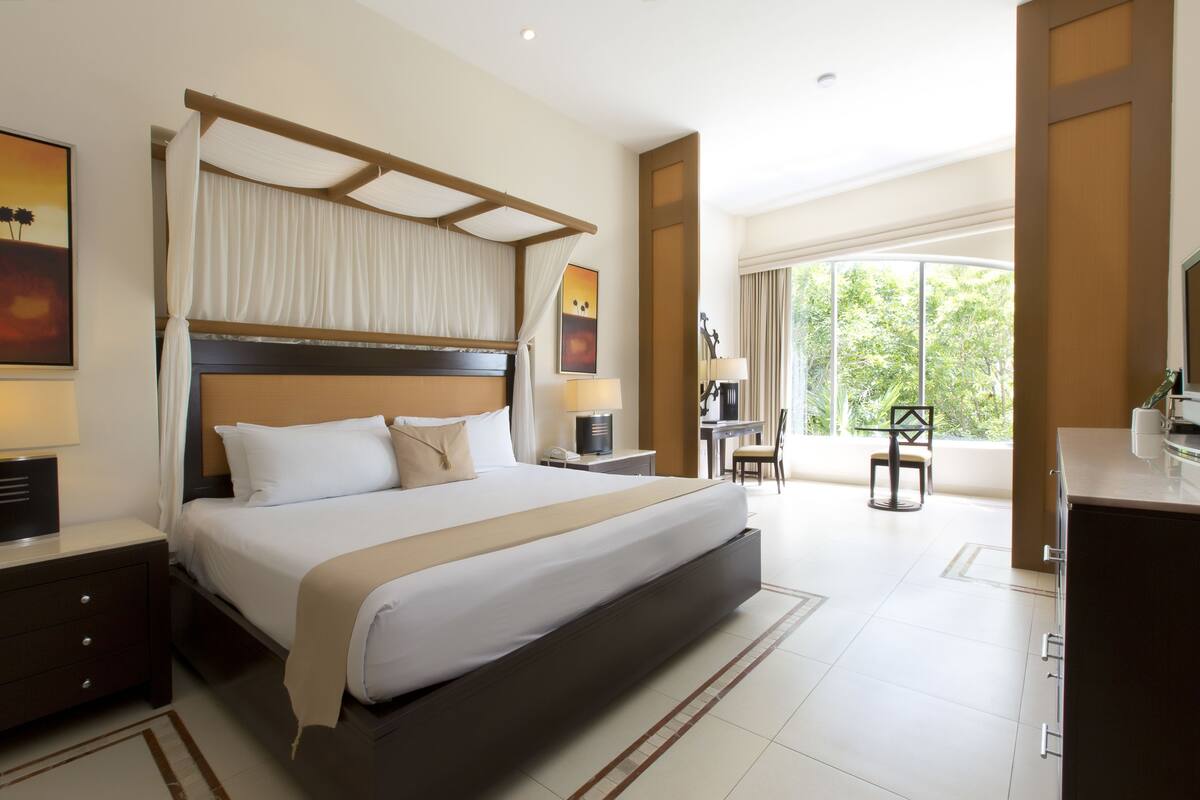 A spacious hotel room with a large bed, a desk, and a view of greenery through the window.