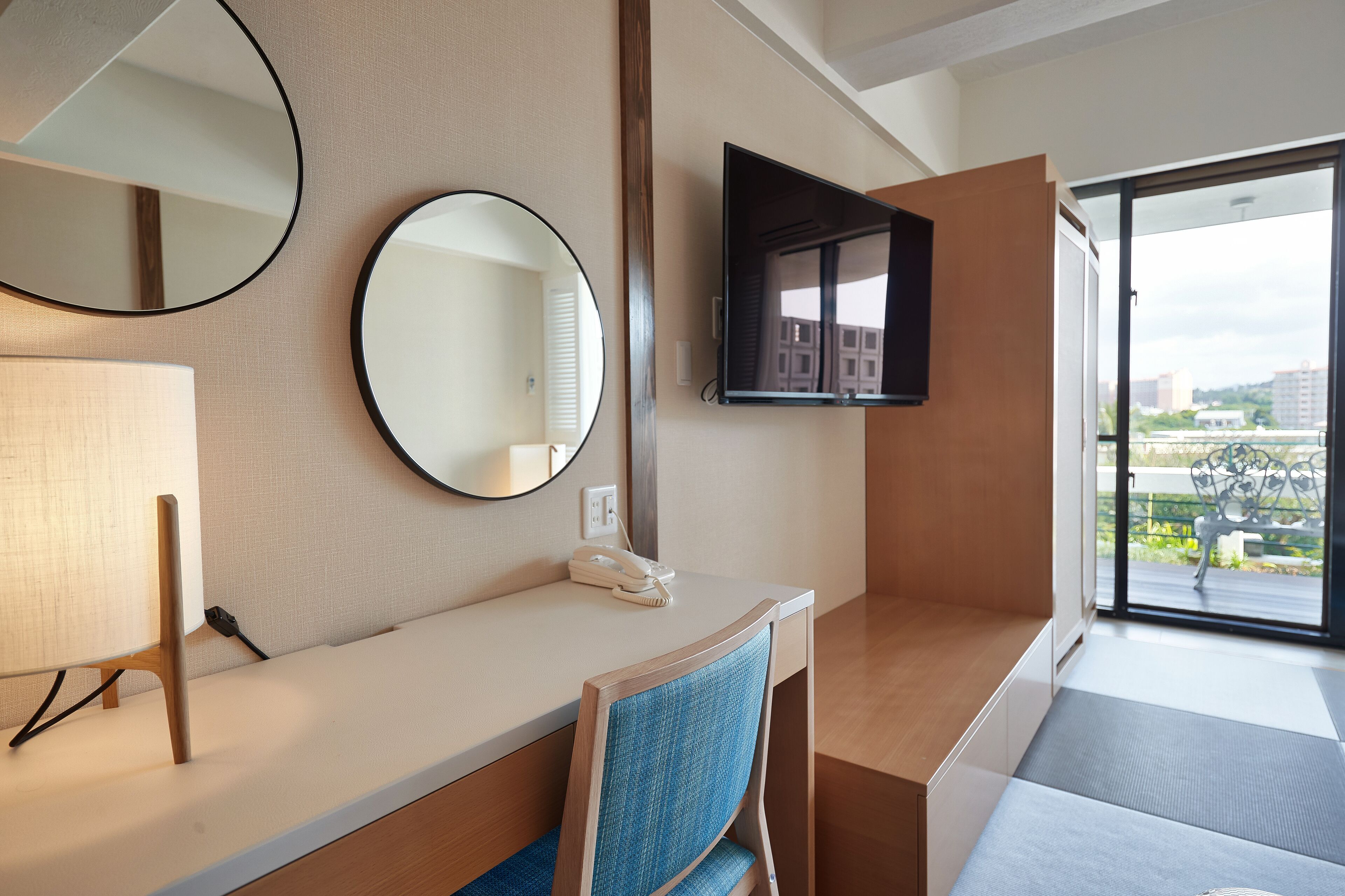 A hotel room with a desk, two round mirrors, a flat-screen TV, a telephone, and a window with a view of buildings and greenery.