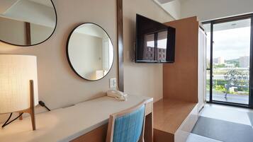 A hotel room with a desk, two round mirrors, a flat-screen TV, a telephone, and a window with a view of buildings and greenery.