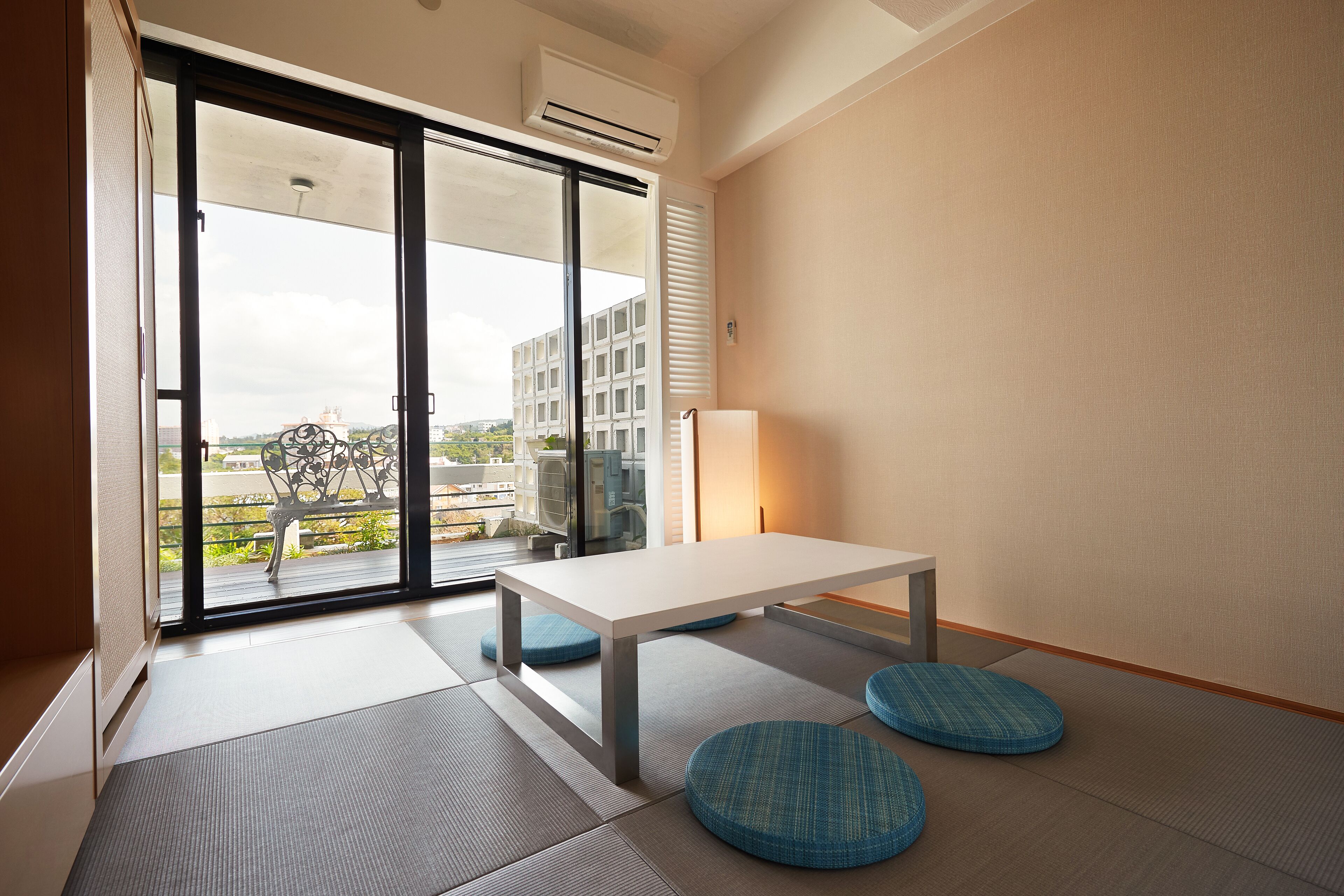Una habitación moderna con una puerta corrediza de vidrio que da a un balcón, una mesa blanca y cojines azules.