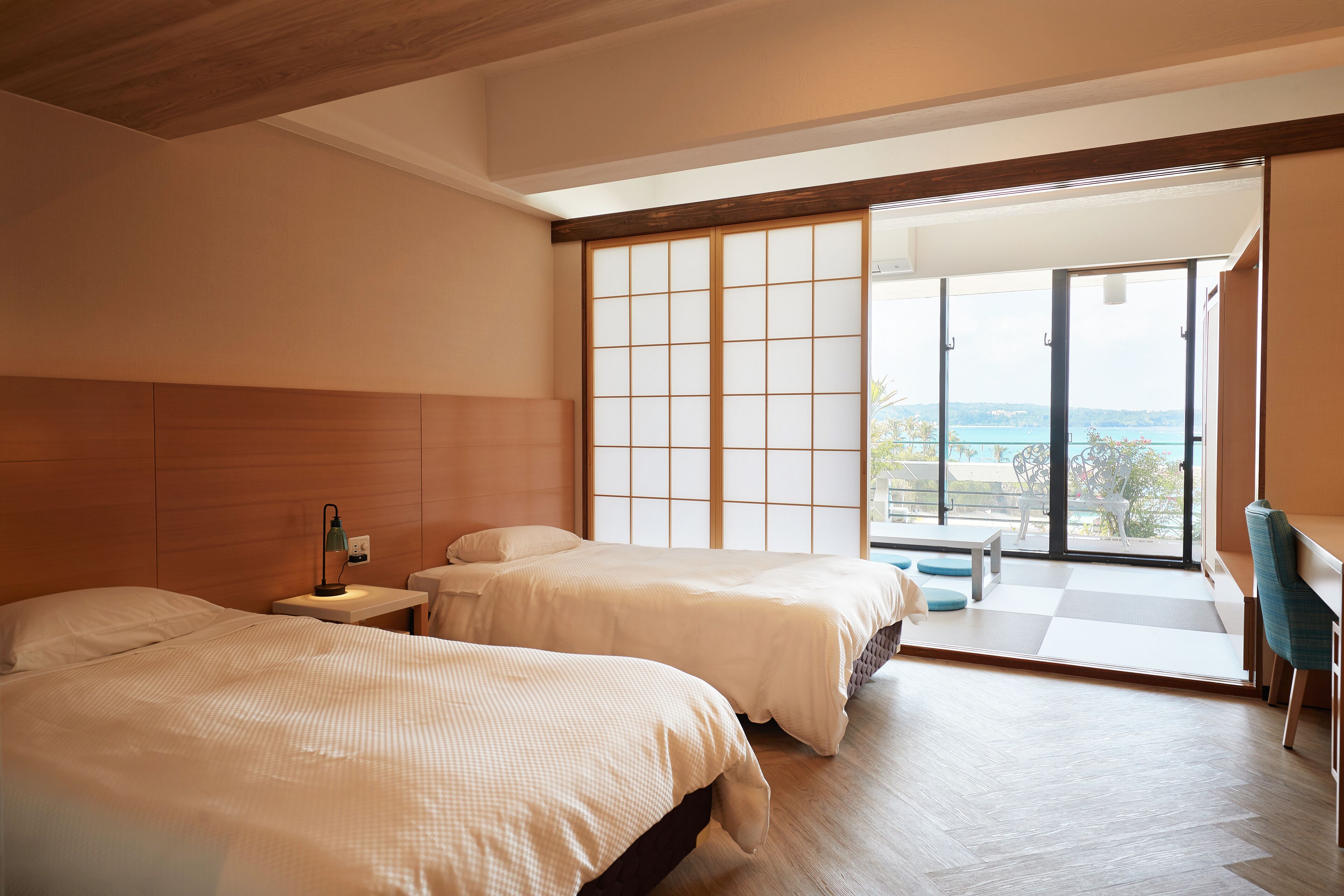 Een moderne hotelkamer met twee bedden, een bureau en een grote schuifdeur die leidt naar een balkon met uitzicht op zee.