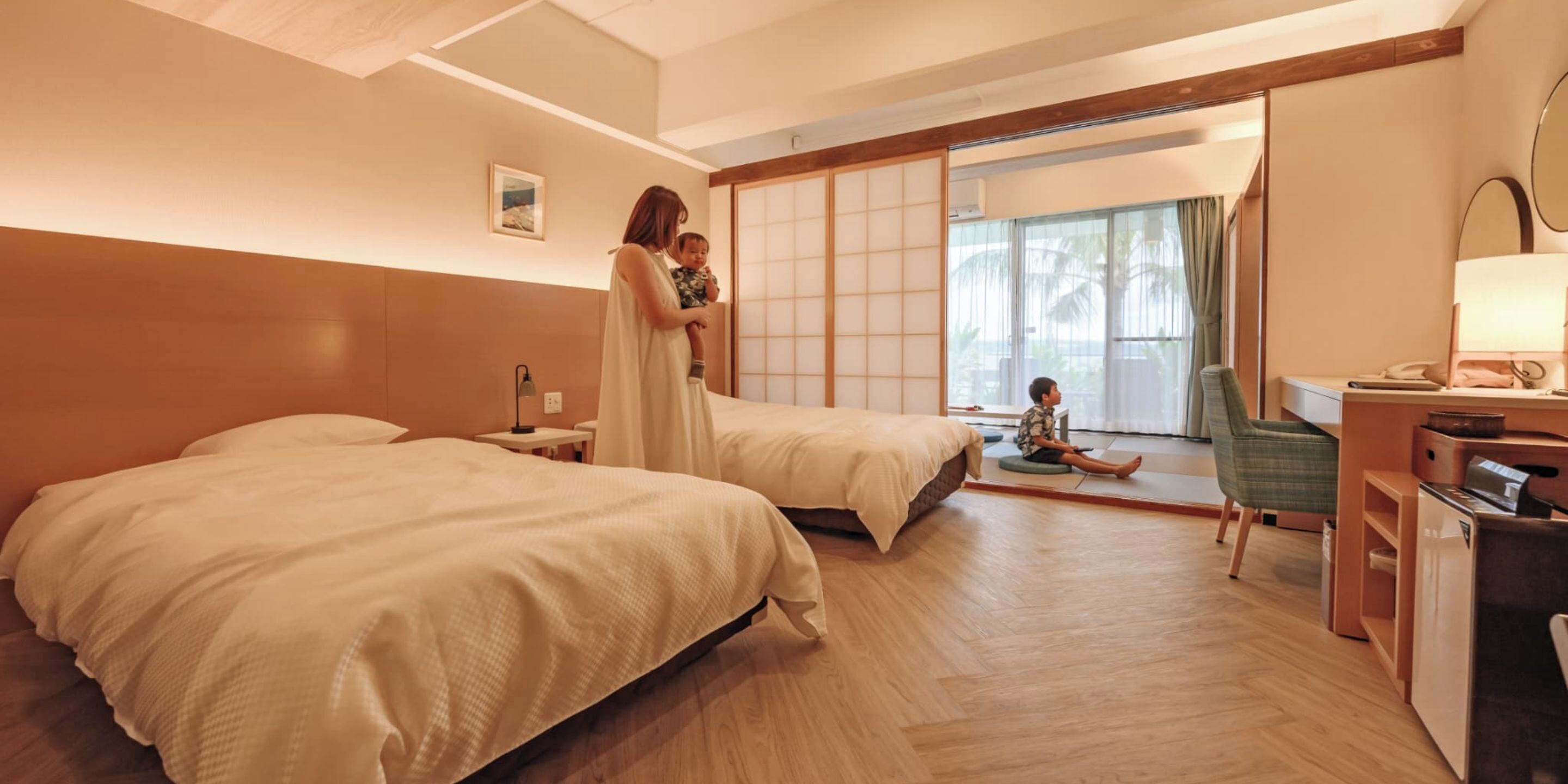 Habitación de hotel con dos camas, un escritorio con lámpara, una silla y vistas a una puerta corredera que da a un balcón.