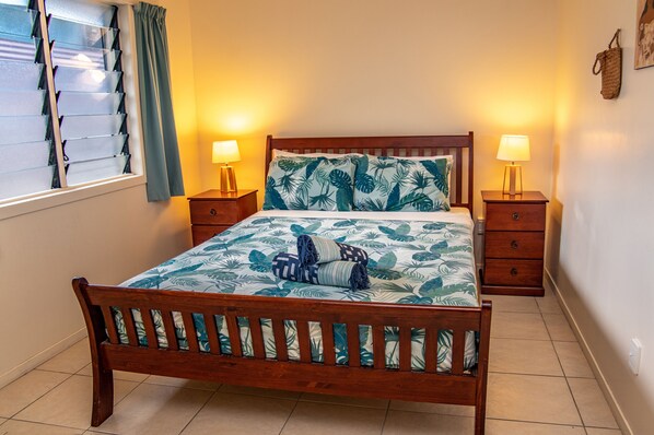 A wooden bed with a tropical leaf-patterned bedspread and pillows, flanked by two bedside tables with lamps, in a room with a window and a wall-mounted bag. - Coral Sands Apartments (Rarotonga)