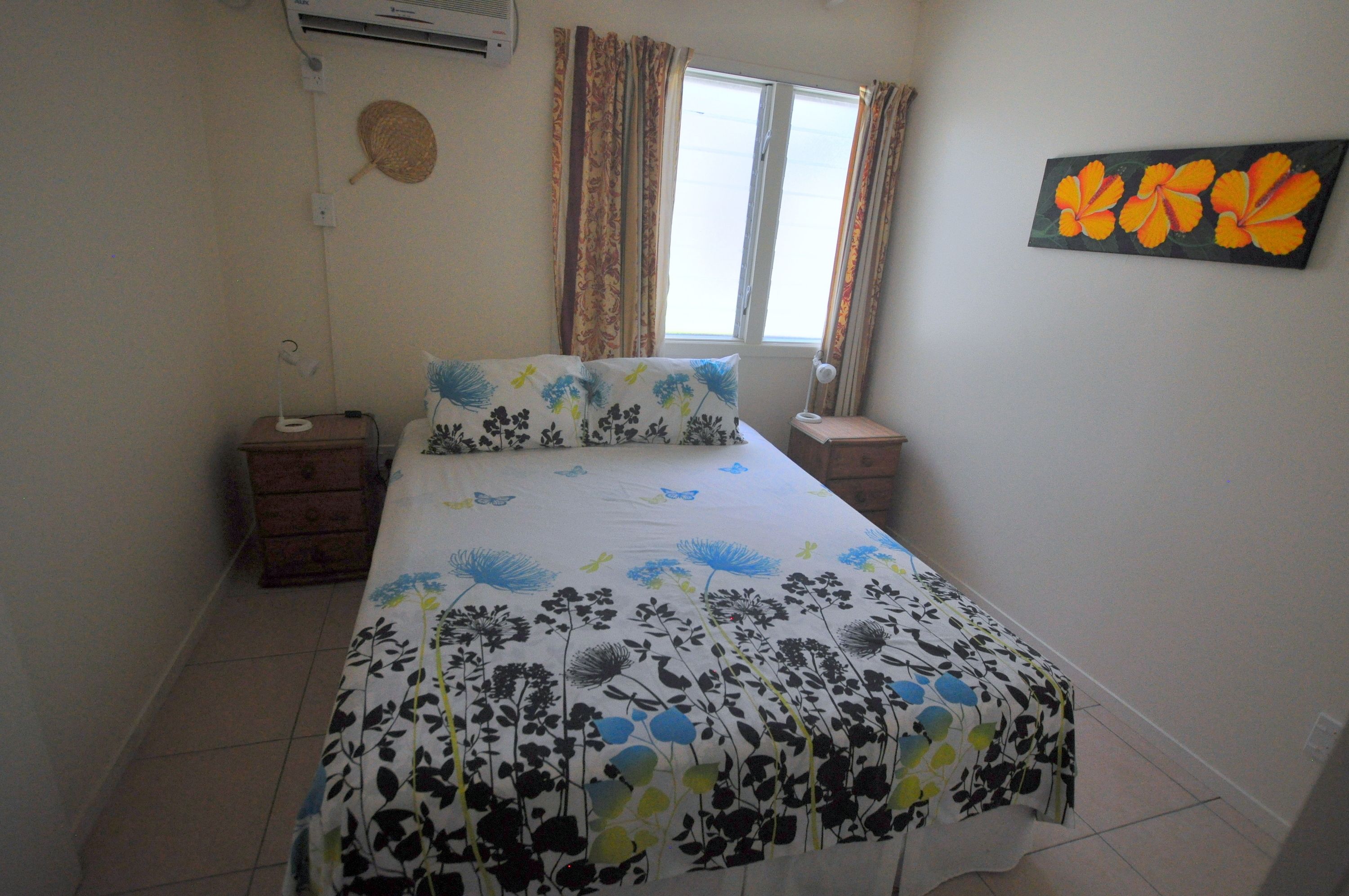 A bedroom with a bed, a nightstand, a lamp, a wall-mounted air conditioner, and a window with curtains.