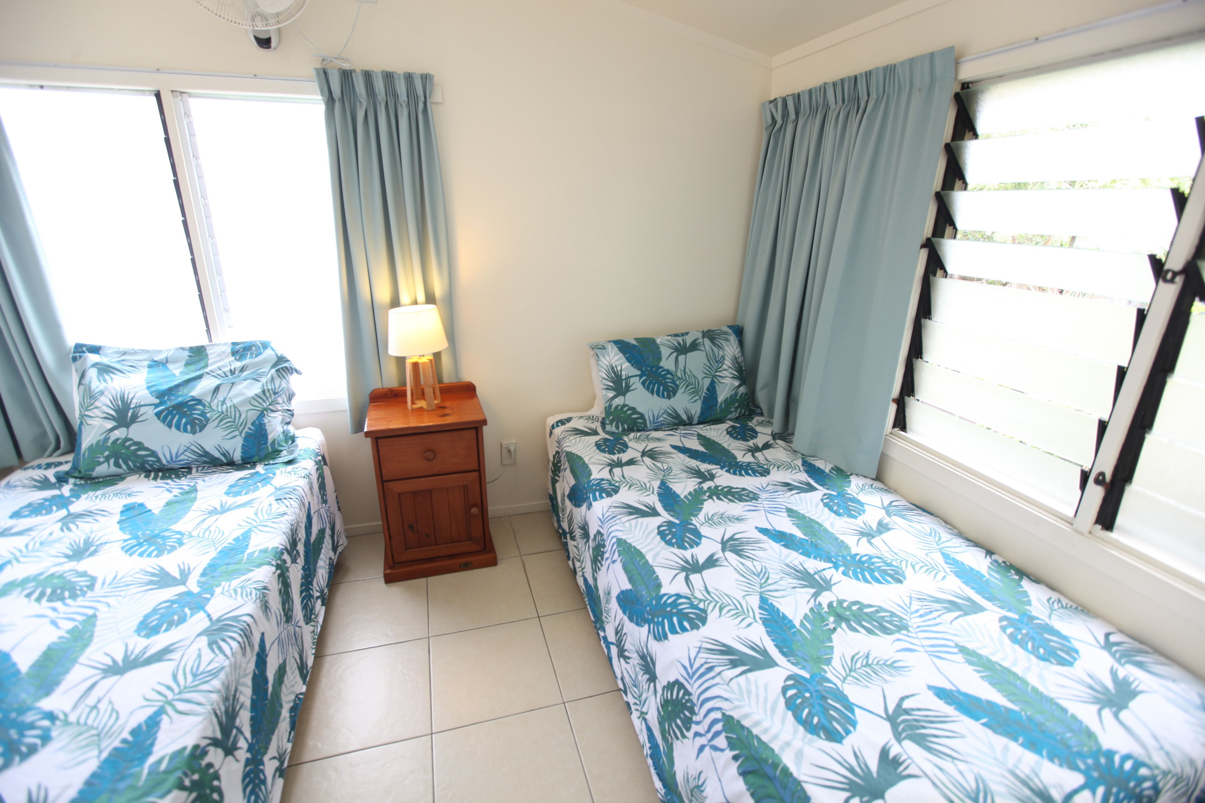 A room with two beds, a nightstand, a lamp, and a window with curtains.