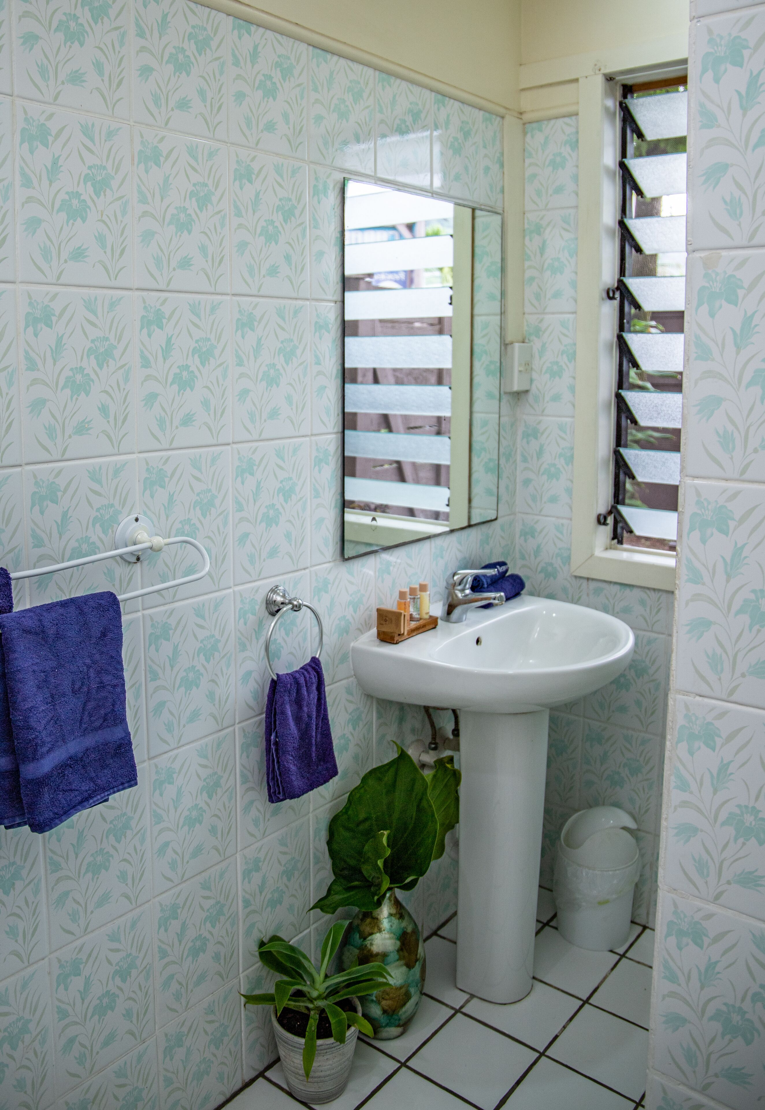 Een badkamer met patroontegels, een wastafel, een spiegel, een raam met zonwering en een plant in een pot.