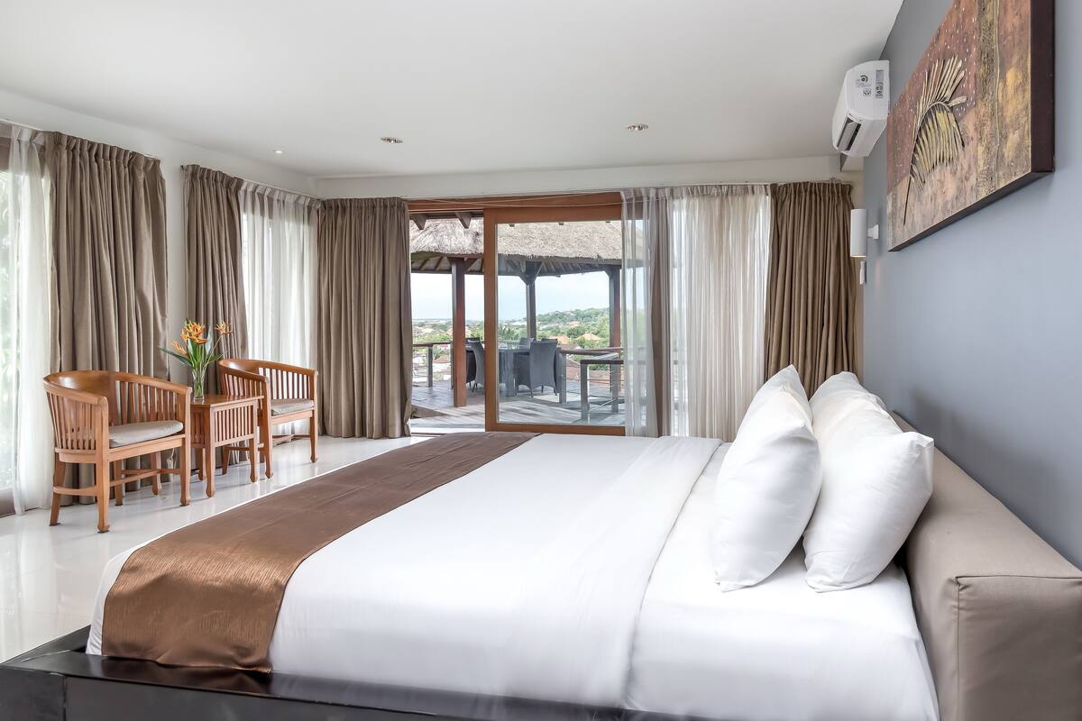 Habitación de hotel con una cama grande, zona de estar con sillas, mesa de comedor y vistas al exterior por un ventanal.