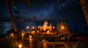 A nighttime outdoor dining area with a table set for two, surrounded by plants and candles.