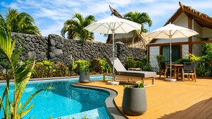 A swimming pool with lounge chairs and umbrellas, surrounded by tropical plants and a stone wall. - Aitutaki Escape (Aitutaki)