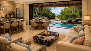 A modern kitchen with a built-in refrigerator, a dining area with a round table and chairs, a poolside seating area with a sofa and lounge chairs, and a view of the ocean. - Aitutaki Escape (Aitutaki)