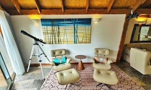 A room with a telescope, two armchairs, a round table, and a large painting of trees.