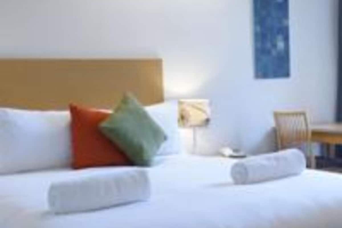 A hotel room with a bed, pillows, a chair, a table, and a wall-mounted lamp.