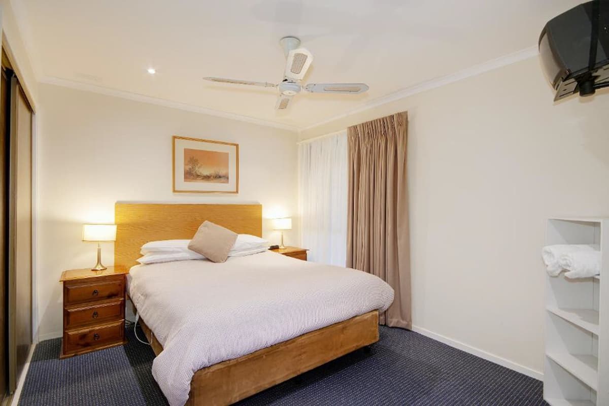 A hotel room with a bed, two bedside tables with lamps, a ceiling fan, a framed picture on the wall, and a window with curtains.