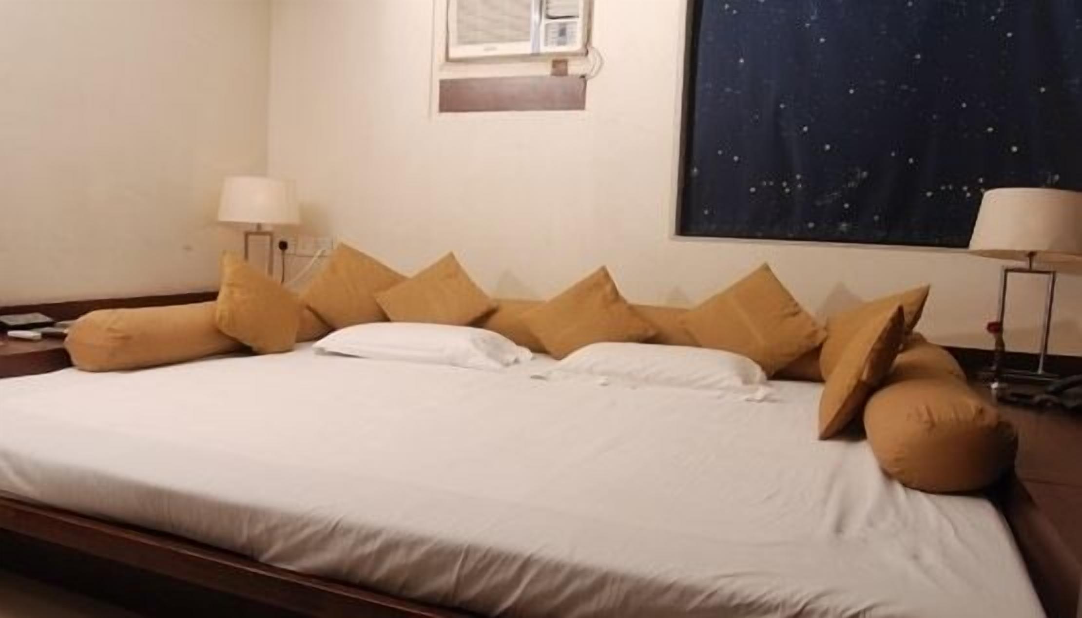 Una cama bien hecha con sábanas blancas y varios almohadones color arena, una mesita de noche con una lámpara y un cuadro grande en la pared con motivos de estrellas.