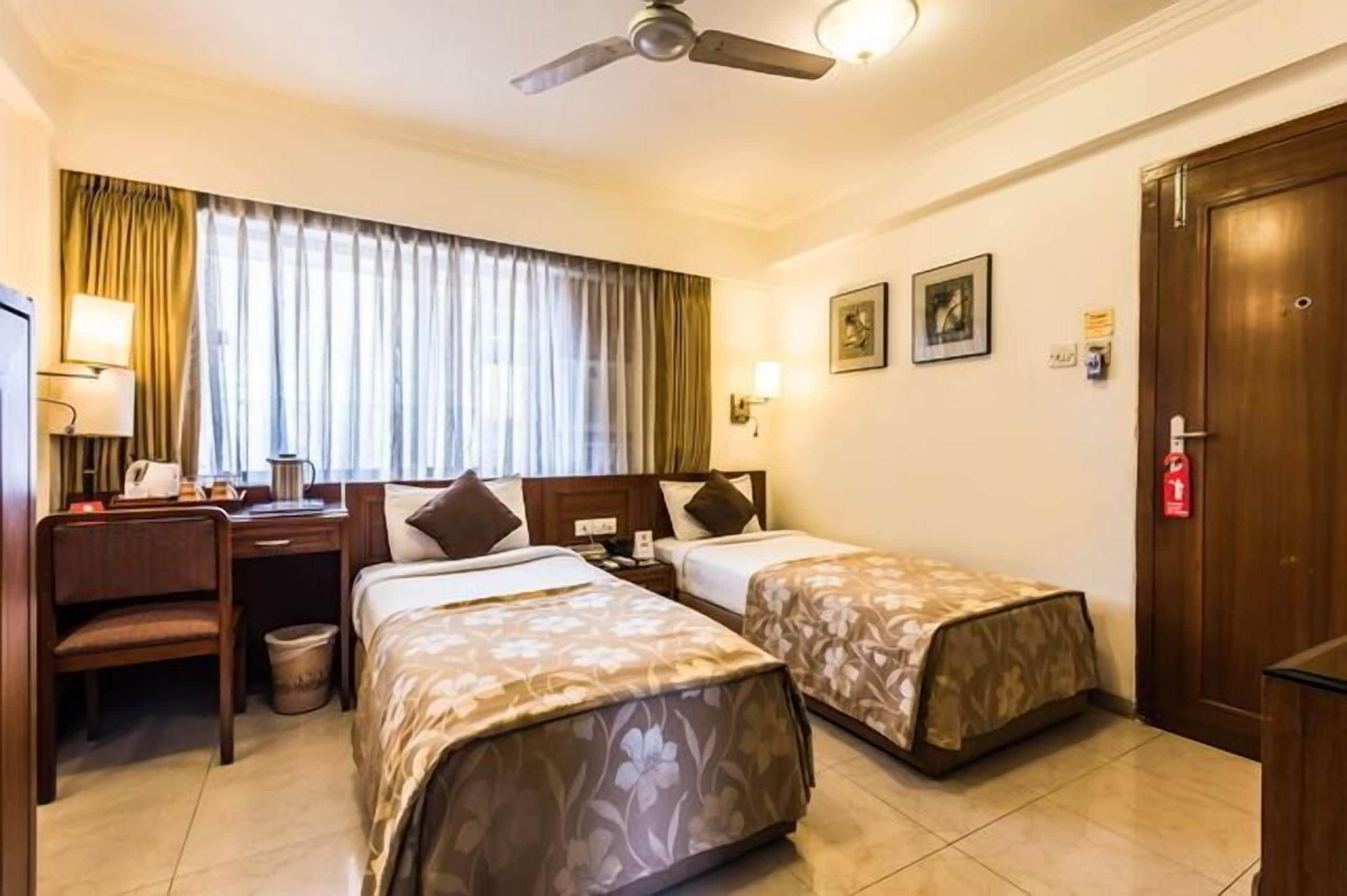 A hotel room with two beds, a desk, a chair, a ceiling fan, and a window with curtains.
