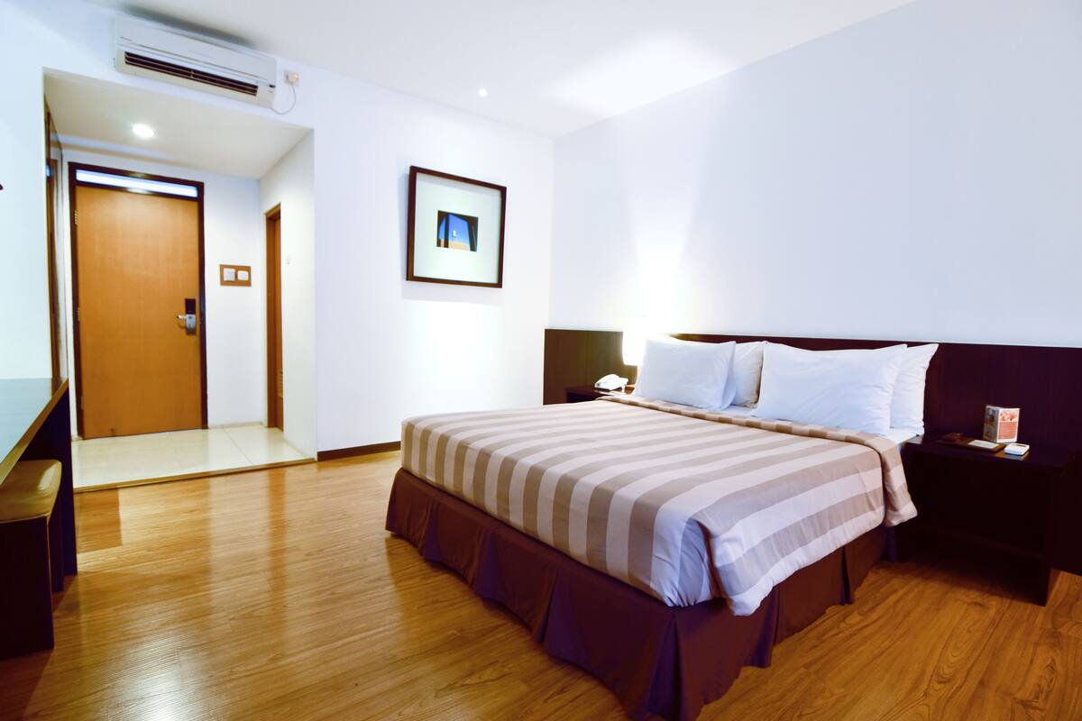 Una habitación de hotel con piso de madera, una cama con ropa de cama a rayas, una mesita de noche con una lámpara y un cuadro enmarcado en la pared.