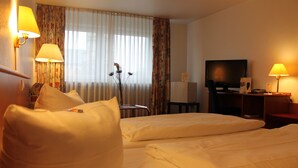 A hotel room with two beds, a desk, a television, and lamps. - City Hotel Essen (Essen)