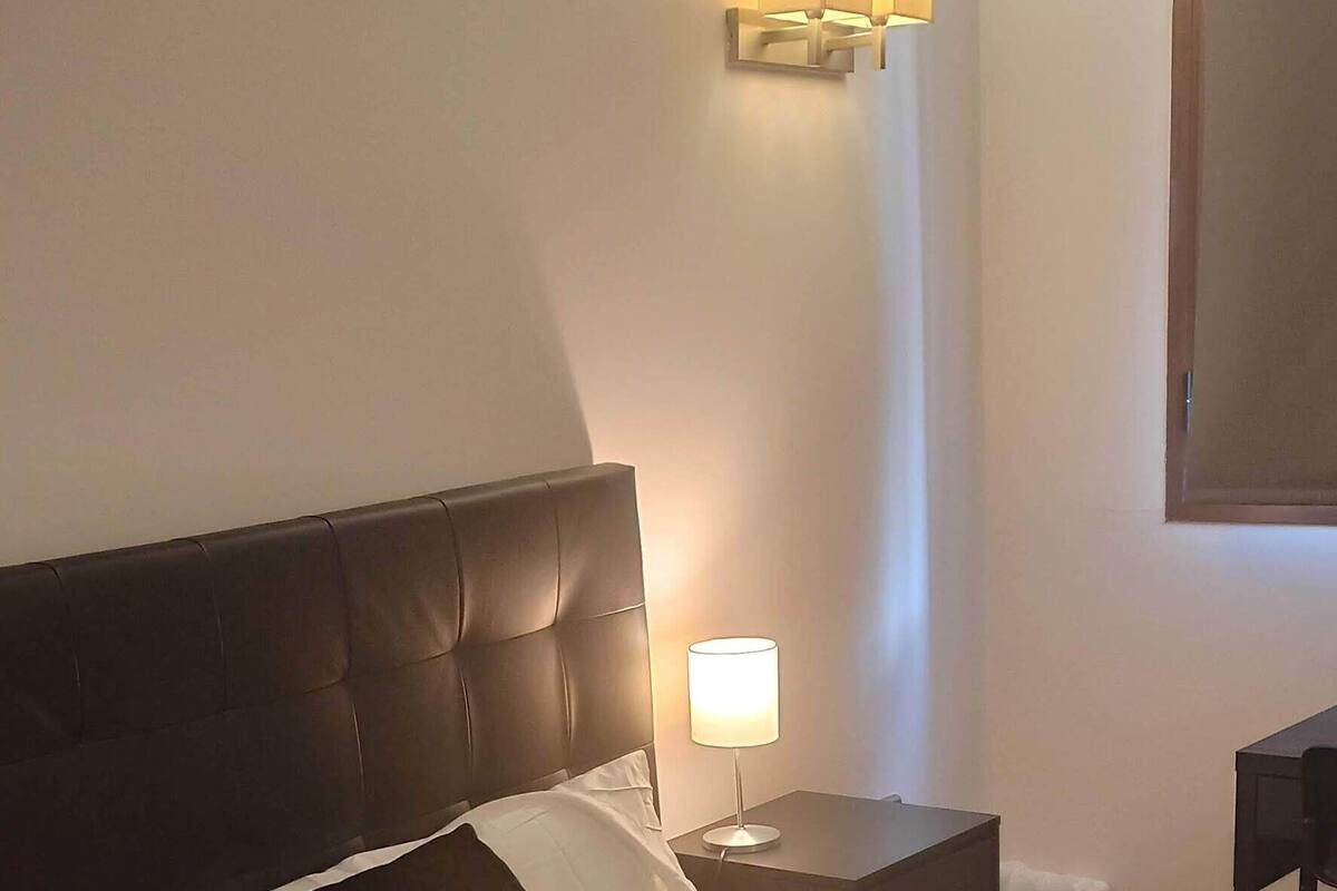 A neatly made bed with a dark headboard, a bedside lamp, and a wall-mounted light fixture.
