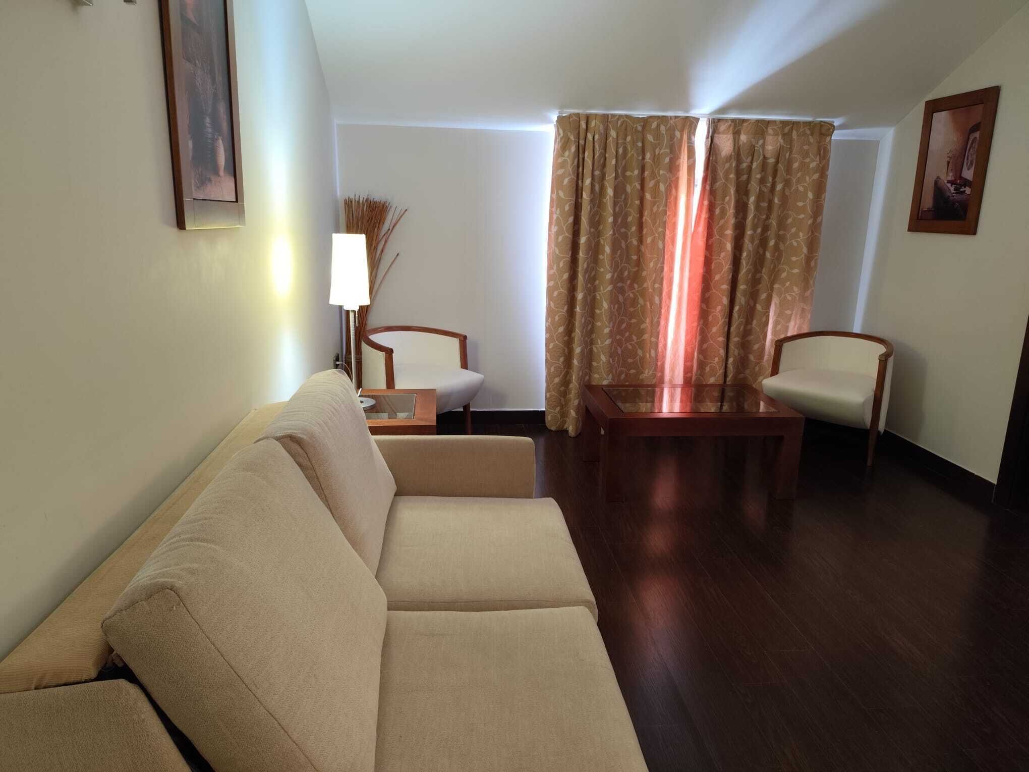 A living room with a sofa, two armchairs, a coffee table, a lamp, and a large window with curtains.