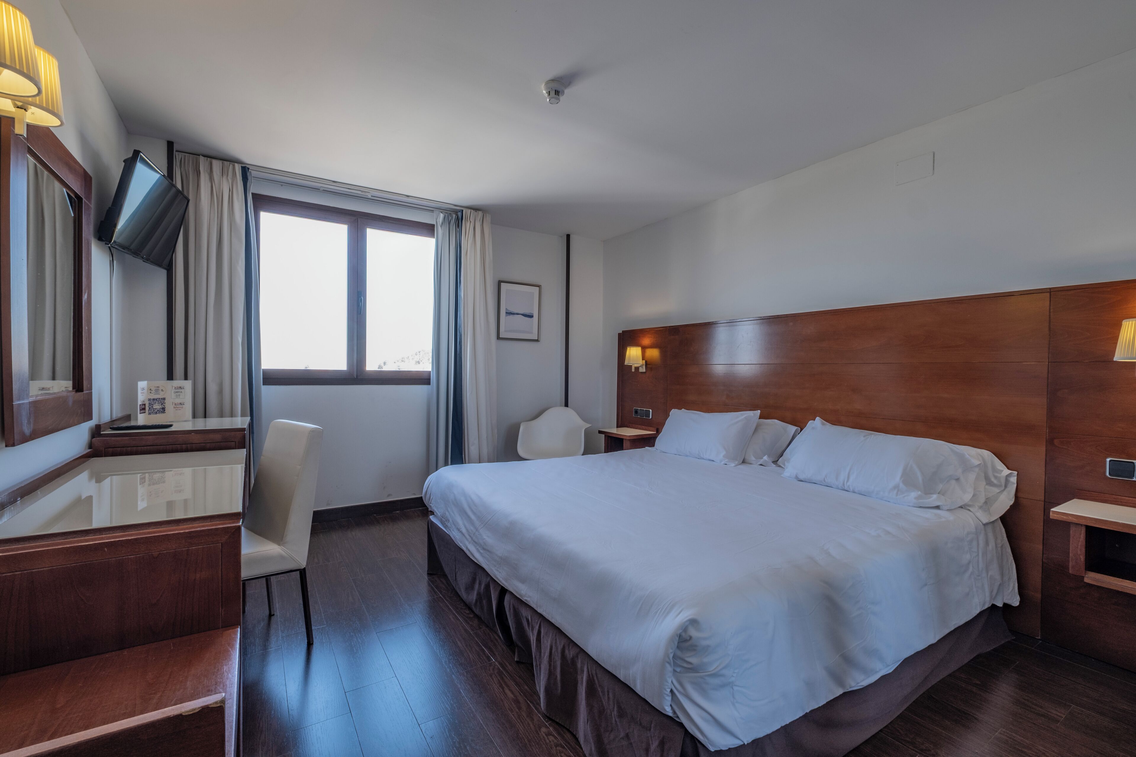 A hotel room with a large bed, a desk with a chair, a TV mounted on the wall, and a window with curtains.