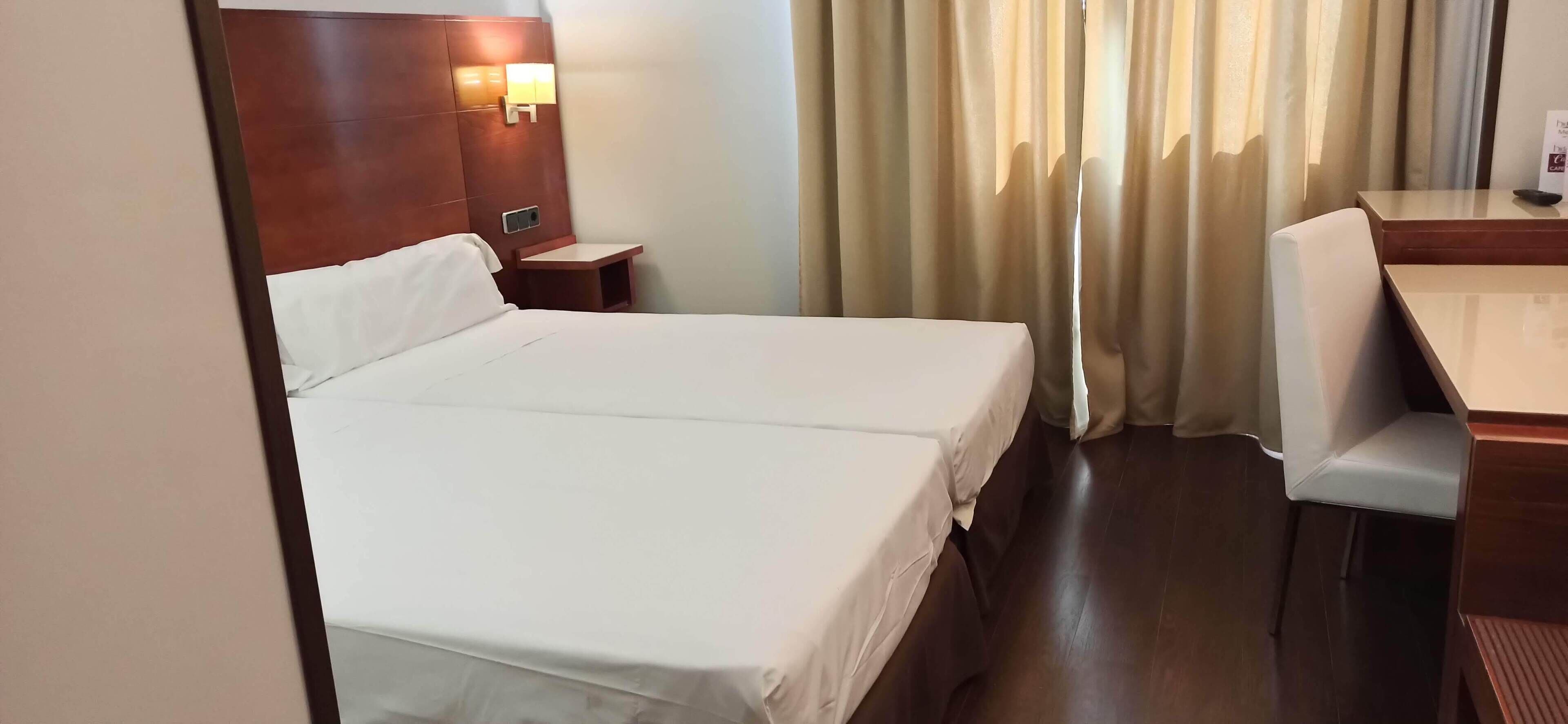 A hotel room with a single bed, a bedside table, a chair, and a desk with a lamp.
