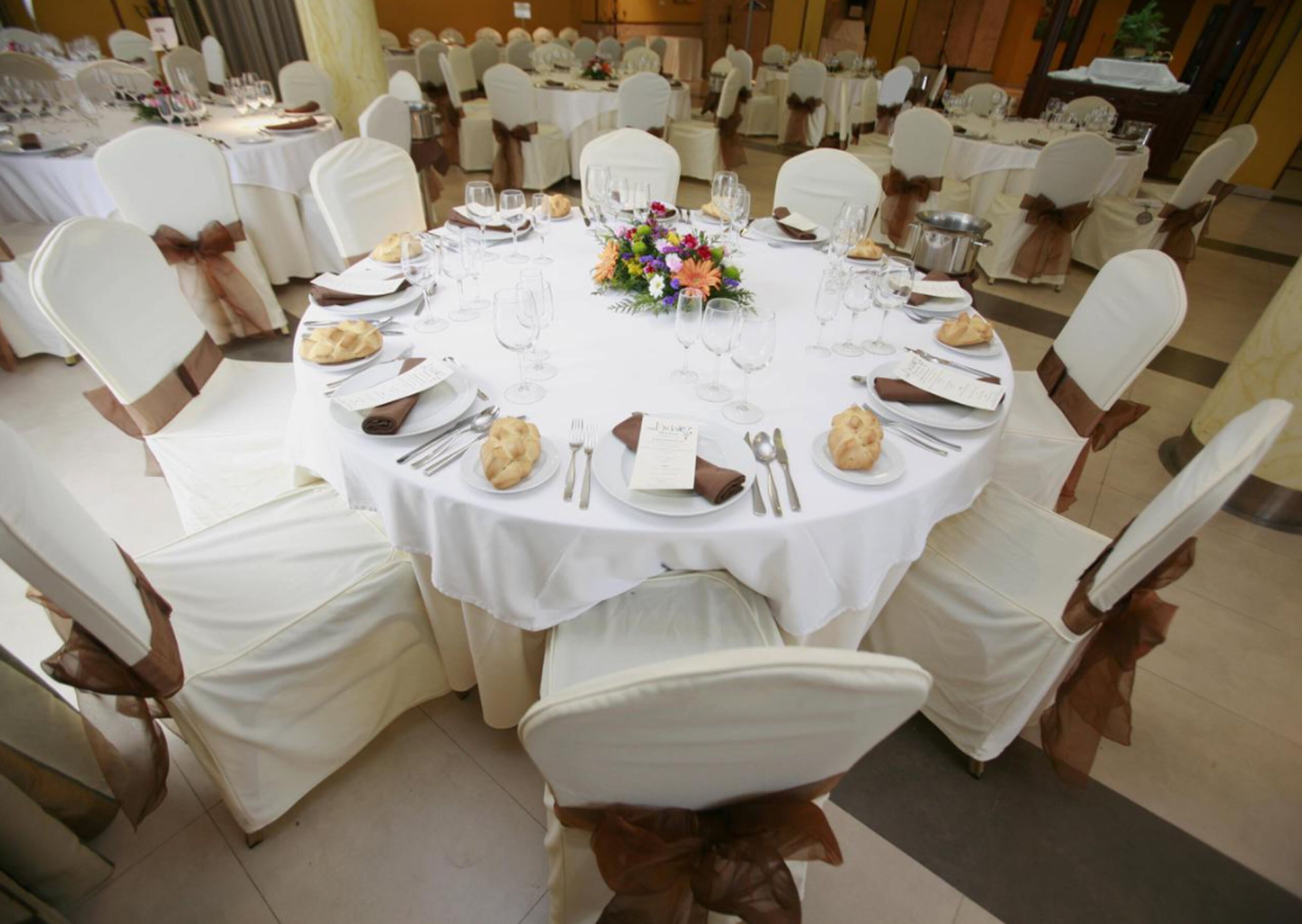 Une table de salle à manger ronde, recouverte d’une nappe blanche, entourée de chaises blanches ornées de rubans marron, préparée pour un repas formel.